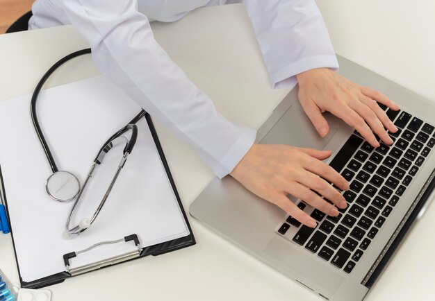 Cropped shot of female doctor hands working with laptop at desk with medical tools and clipboard