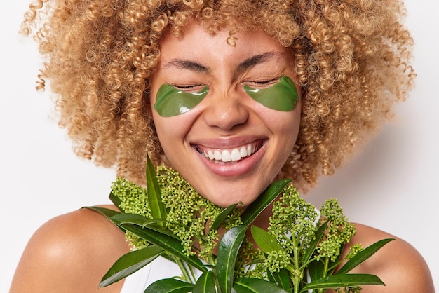 Cropped shot of overjoyed curly haired woman smiles broadly keeps eyes closed giggles positively holds green fresh plant applies hydrogel pads isolated over white background. Eye skin concept