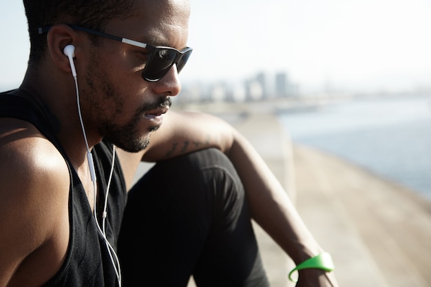 Free photo cropped shot of pensive young handsome jogger in sunglasses sitting on top of stone stairs, dressed in black sportswear, looking sad and serious, listening to music on his smart phone , having rest