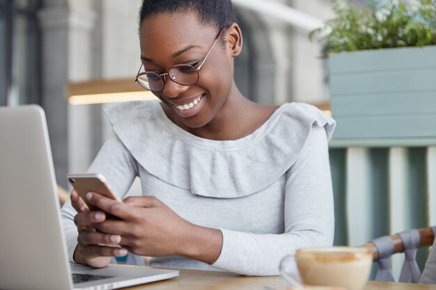 Cropped shot of pleased female copywriter reads positive information on smart phone, sits in front of opened laptop computer drinks aromatic coffee.