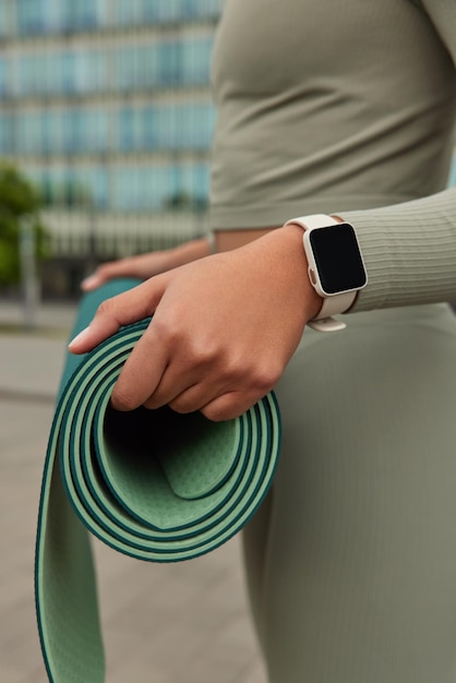 Free photo cropped shot of unrecognizable woman in activewear carries rolled up karemat gets ready for workout wears smartwatch and tracksuit poses against blurred background. sporty lifestyle concept.