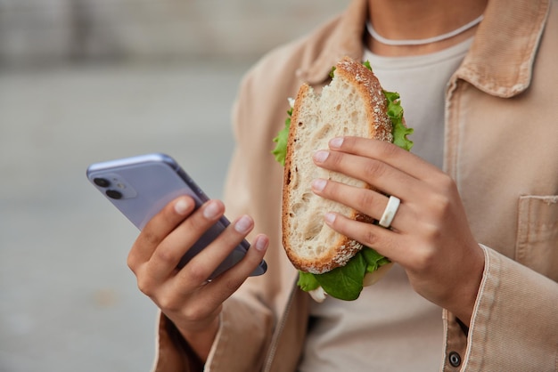 Free Photo cropped shot of unrecognizable woman eats delicious sandwich and uses mobile phone for texting messages dressed in stylish clothes poses outdoors against blurred background. fast food concept