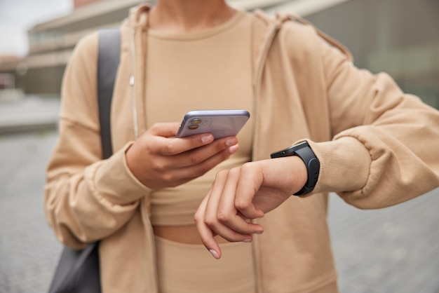 Free photo cropped shot of unrecognizable woman holds modern mobile phone checks results of fitness traning dressed in sportswear carries fitness mat poses outdoors. people sport and technology concept