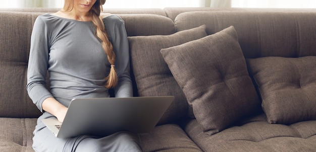 Free Photo cropped shot of young blond woman with braid working on a laptop sitting on the comfortable dark sofa at home, backlit warm light. freelance or lifestyle concept.