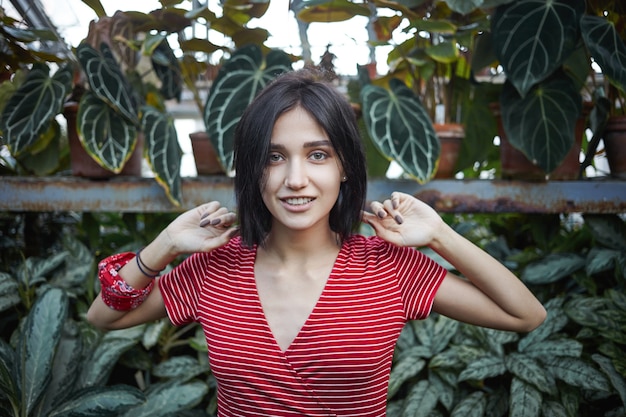 Free photo cropped view of beautiful young european female with dark bib hairstyle relaxing outdoors, walking in jungle surrounded with exotic plants, feeling happy and carefree, smiling joyfully at camera