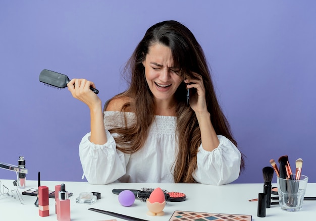 Free photo crying beautiful girl sits at table with makeup tools holds hair comb talking on phone looking down isolated on purple wall