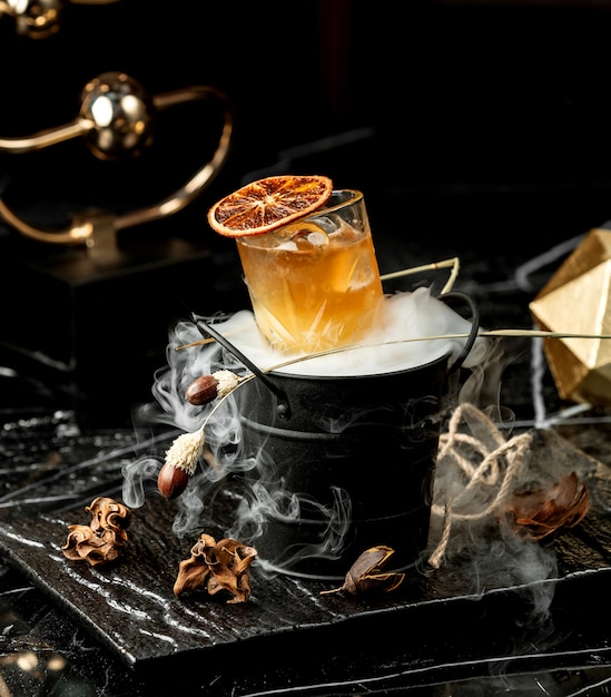 crystal glass of cold cocktail garnished with dried orange slice served in smoked bucket