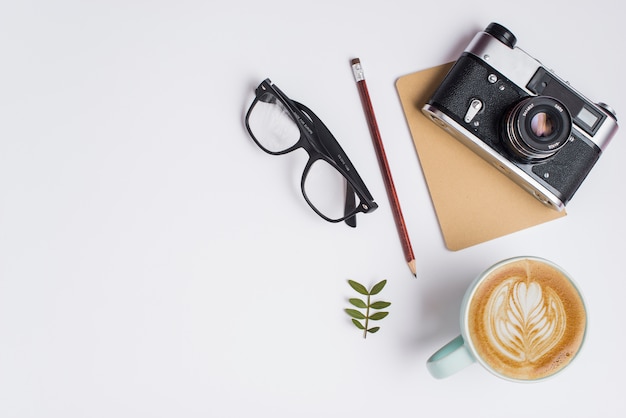 Free photo cup of coffee latte; pencil; eyeglasses and vintage camera on white background