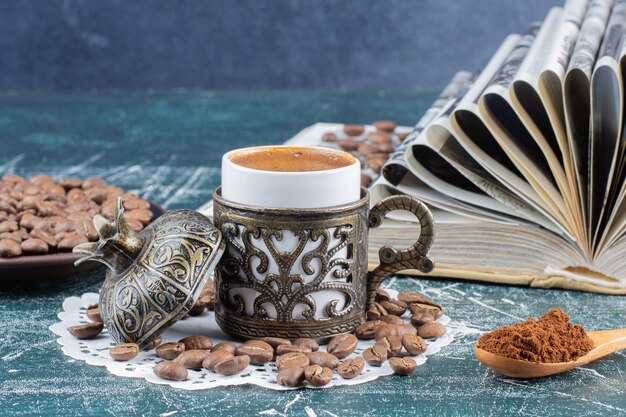 Cup of foamy coffee, plate of coffee beans and book on marble table.