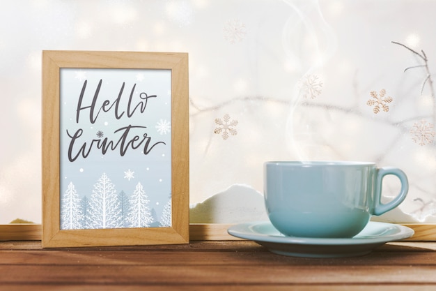 Cup near frame with hello winter title on wood table near bank of snow