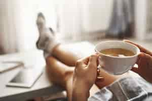 Free photo cup of tea and chill. woman lying on couch, holding legs on coffee table, drinking hot coffee and enjoying morning, being in dreamy and relaxed mood. girl in oversized shirt takes break at home