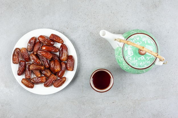 Free photo a cup of tea and an ornate teapot next to a plate of fresh dates on marble background. high quality illustration