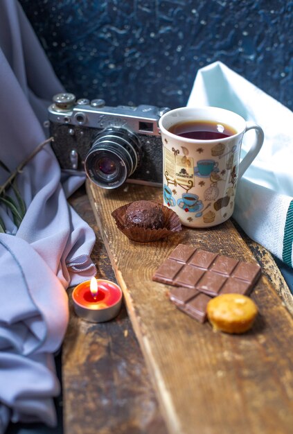 A cup of tea with chocolate bar and muffins on a piece of wood.