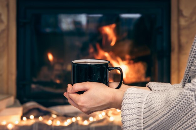 A cup with a hot drink in female hands on a blurred fireplace background