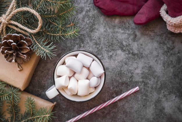 Cup with marshmallows near plastic tube, fir twigs and present boxes 