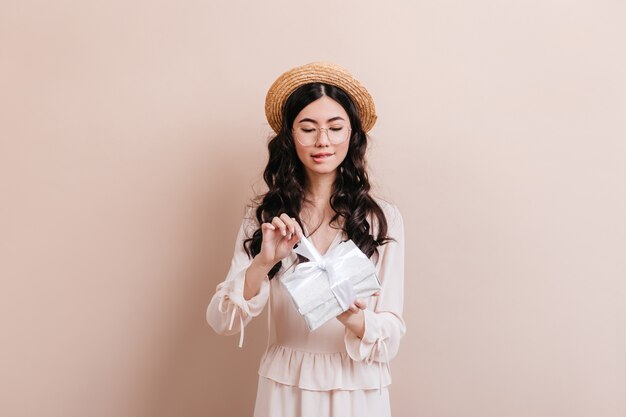 Curious asian woman opening birthday present. Cute chinese female model holding gift on beige background.