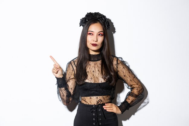 Curious attractive asian woman in witch costume, pointing finger upper left corner, looking interested in halloween promo, standing over white wall and thinking