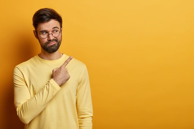 Curious bearded man points ay copy space on yellow wall, demonstrates important information, found solution or answer on question, says his suggestion, wears yellow jumper, round spectacles