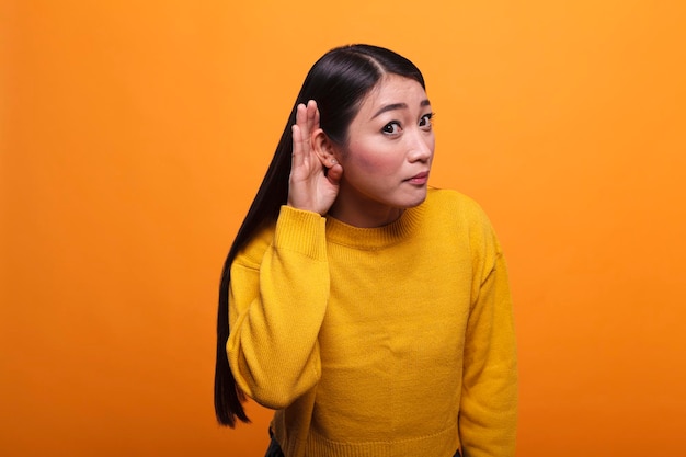 Curious brunette asian woman eavesdropping interesting gossip and secret rumours about her. Intrigued serious person with hand at ear overhearing people gossiping with interest.