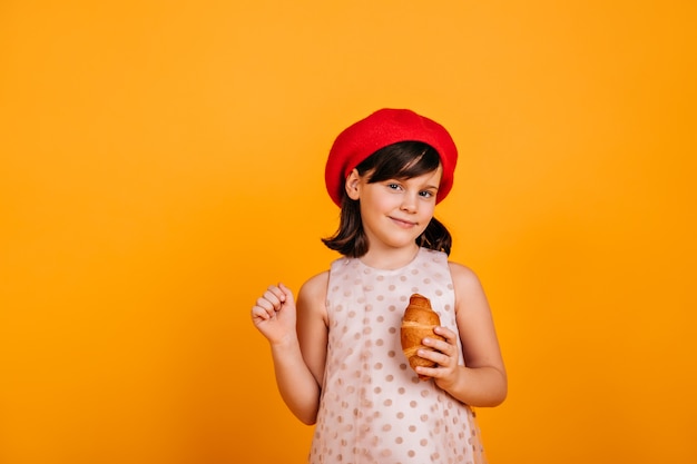 Curious brunette kid posing on yellow wall.  preteen girl eating croissant.