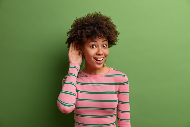 Free Photo curious cheerful ethnic young woman holds hand near ear, eavesdrops rumors, overhears interesting conversation, likes to know every news, wears striped jumper, poses over vivid green  wall