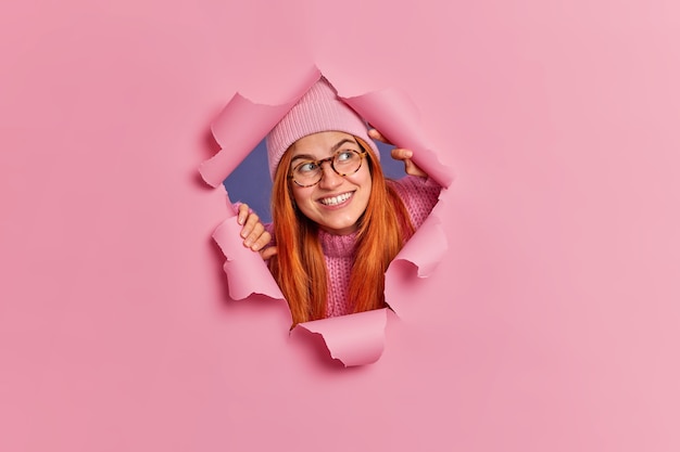 Free Photo curious cheerful redhead woman concentrated aside sees something very pleasant smiles broadly wears hat and spectacles.