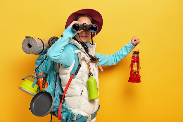 Free photo curious female backpacker explores tourist destination, uses binoculars, dressed in active wear, holds kerosene lamp carries travelling items with rucksack