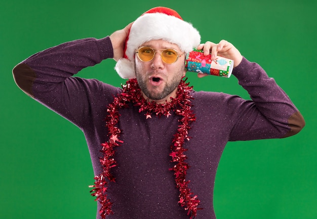 Curious middle-aged man wearing santa hat and tinsel garland around neck with glasses holding plastic christmas cup next to ear listening to secrets 