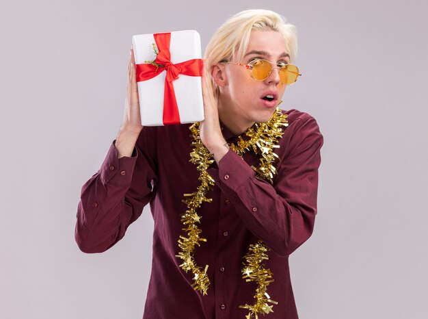 Curious young blonde man wearing glasses with tinsel garland around neck holding gift package near head looking at camera isolated on white background