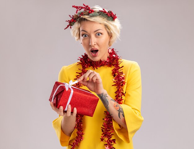 Curious young blonde woman wearing christmas head wreath and tinsel garland around neck holding christmas gift package looking at camera grabbing ribbon isolated on white background