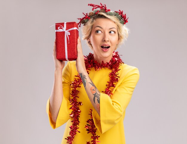 Curious young blonde woman wearing christmas head wreath and tinsel garland around neck holding christmas gift package near head looking at it isolated on white background