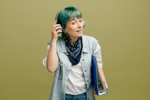 Curious young female student wearing headphones and bandana on neck holding folder looking at camera opening one ear trying to hear someone through loud sound of music