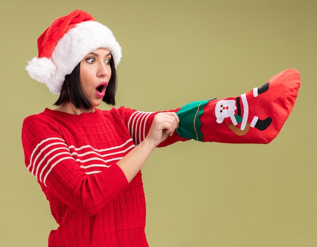 Free photo curious young girl wearing santa hat holding christmas stocking looking at it putting hand inside it isolated on olive green background