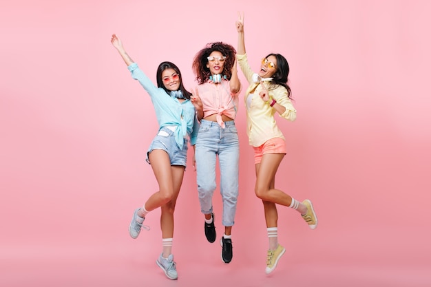 Curly african woman in jeans jumping while posing with international university friends. Tanned latin girl in yellow shirt dancing and having fun with other ladies.