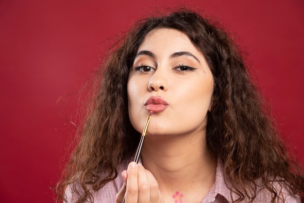 Curly artist looking applying her lipstick with paintbrush.