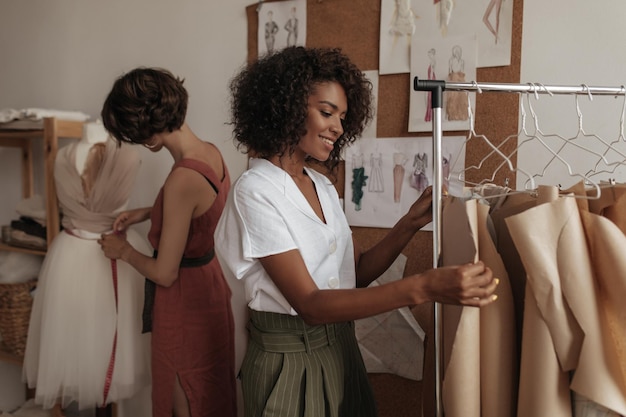 Curly attractive darkskinned woman in white blouse and khaki shorts works as fashion designer Shorthaired lady designs dress