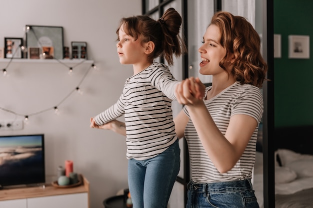 Curly beautiful woman in striped T-shirt raised her daughter high and playing with her.