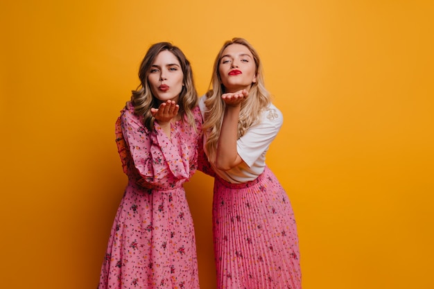 Curly brunette woman in long dress posing with best friend. Pleased caucasian girls sending air kisses on yellow wall.