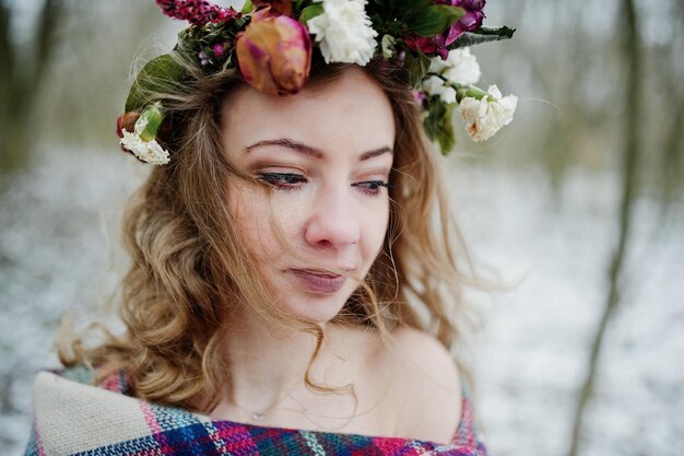 Curly cute blonde girl with wreath in checkered plaid at snowy forest in winter day