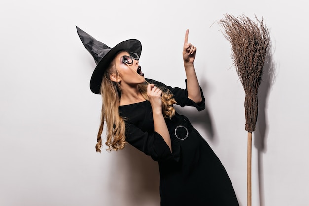 Free Photo curly girl in halloween costume looking up. charming witch in black hat posing with old broom.