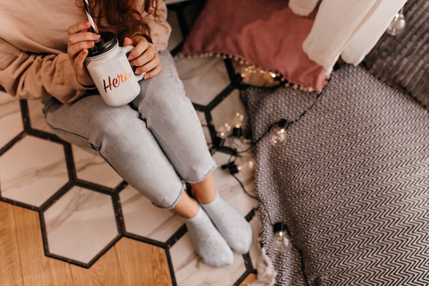 Free photo curly girl in jeans sitting on the floor and drinking hot beverage. indoor overhead portrait of female model with cup of tea.