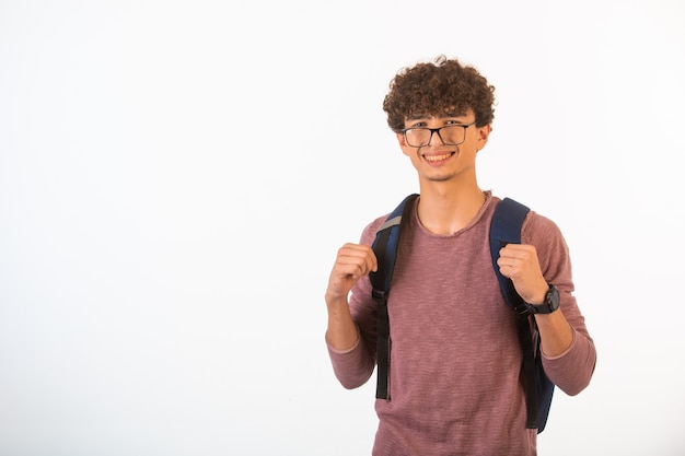 Curly hair boy in optique glasses holding his backpack is confident and motivated, front view. 