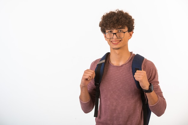 Curly hair boy in optique glasses holding his backpack is happy and joyful, front view.