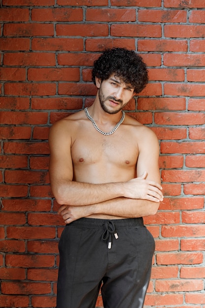 Curly haired half naked man posing to camera against wall.