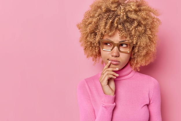 Curly haired young European woman ponders on something keeps hand near mouth considers wears spectacles and casual turtleneck isolated over pink background copy space for your promotional content