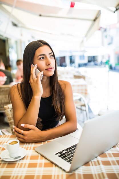 Curly latin girl talking on phone while sitting in outdoor cafe. Pretty female freelancer in glasses chilling in restaurant.