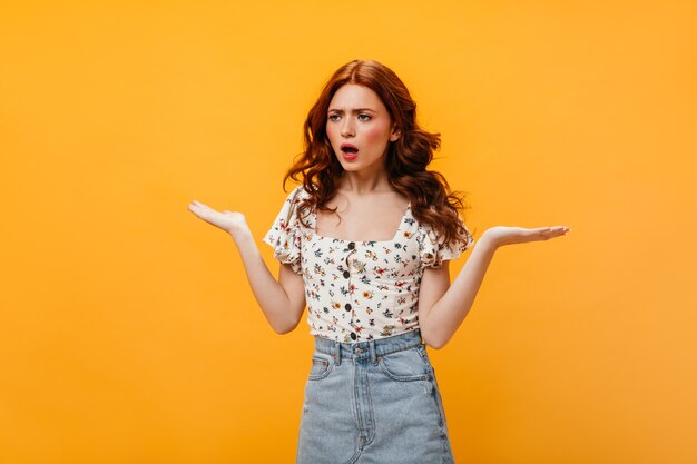 Curly woman in astonishment spreads her hands. Portrait of outraged woman in white top and denim skirt.