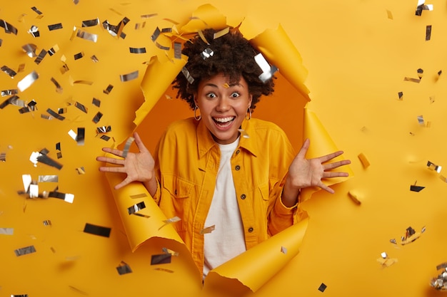 curly woman excited about many sales started at shopping mall, fascinated by awesome news, spreads palms and smiles toothy