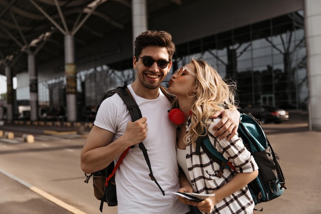 Free photo curly woman kisses her boyfriend near airport attractive girl in plaid shirt and brunette man in white tee poses in good mood and holds backpacks