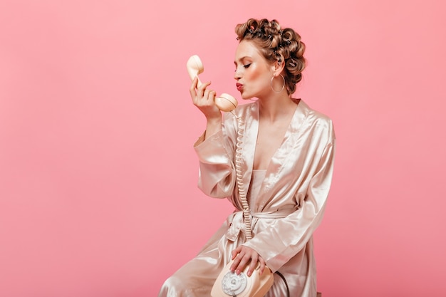 Free photo curly woman in silk home outfit sits on chair on pink wall and kisses phone